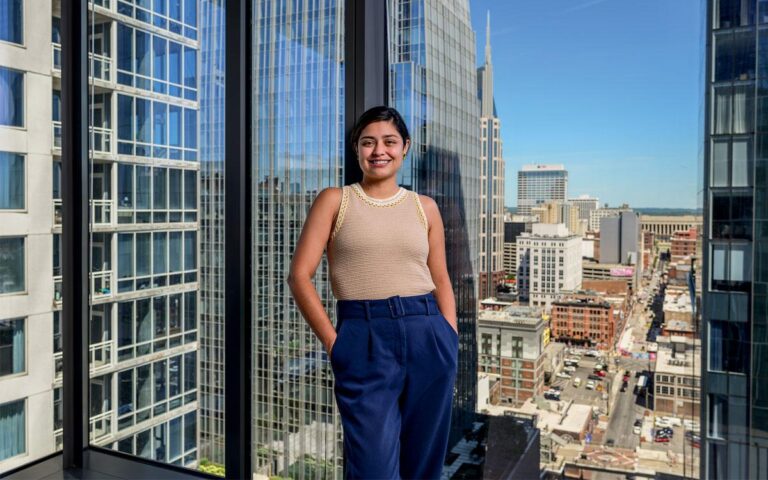 Laura Zapata stands against a glass wall inside a skyscraper with a city in the background