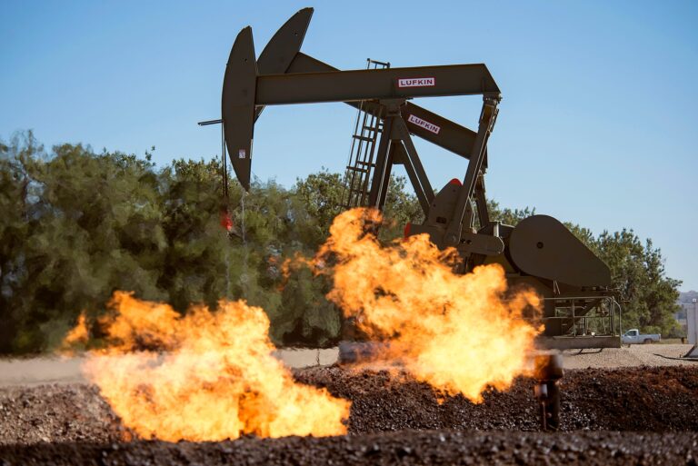 Flames rise out of low pipes in the ground as two jack pumps work in the background under a blue sky