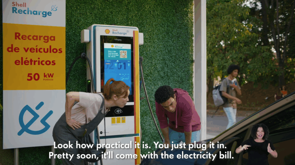 A video still of a Shell ad in Brazil. A man and a woman at a Shell station charging point are about to attach the electric vehicle charging cable to their car. An English subtitle at the bottom of the screen, Look how practical it is. You just plug it in. Pretty soon, it