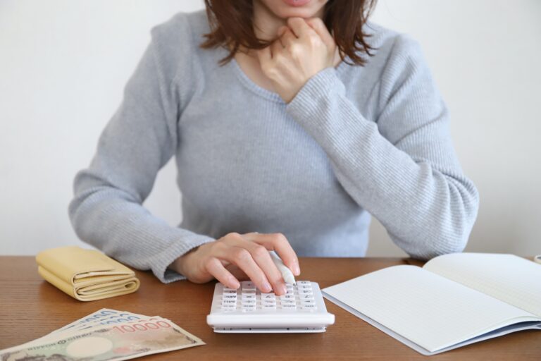 A woman in a gray sweater sits at a desk using a calculator, with cash, a wallet, and an open notebook spread out in front of her.