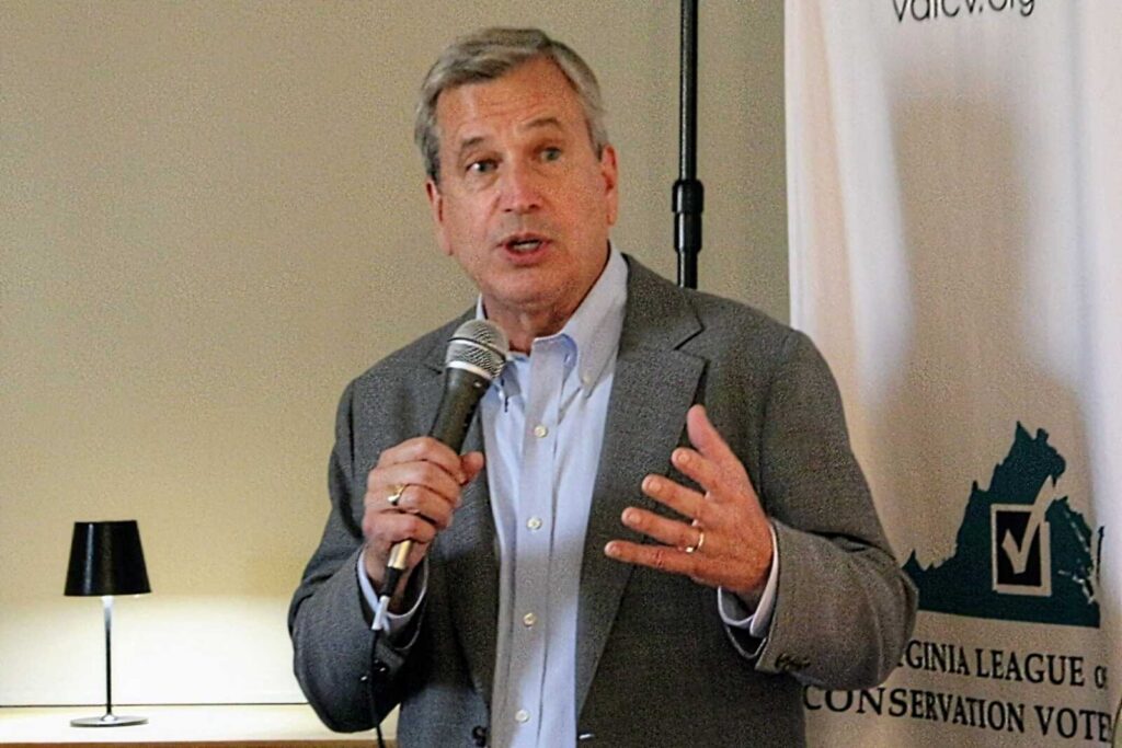 Man in a gray blazer speaks into a handheld microphone at a public event, with a banner in the background.