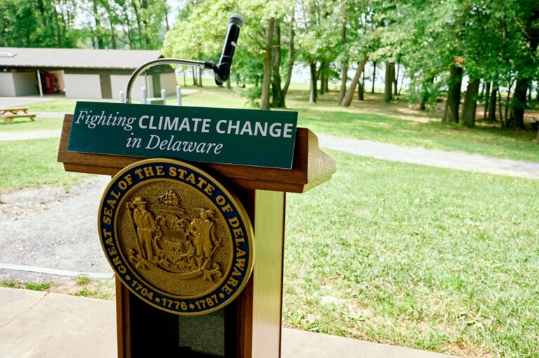 Delawares Climate Action Reshaping the States Future and Economy - Creation Covenant Alliance A podium with the Delaware seal and a sign about fighting climate change sits in front of trees.